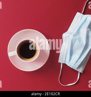 Maschera medica protettiva e tazza di caffè sono su un tavolo rosso. Maschera di procedura da batteri. Il concetto di vita nell'era del coronavirus. Lifestyle Foto Stock