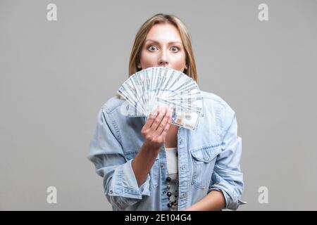 Ritratto di sorpreso giovane donna in denim shirt che spuntavano di dollari ventola, nascondendo la sua metà faccia e guardare la fotocamera con grandi occhi attoniti. interna s Foto Stock