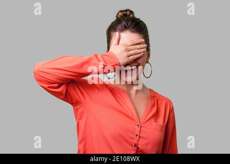 Non si desidera guardare! Ritratto di positivo donna elegante con bun hairstyle, grandi orecchini e in rosso camicetta in piedi che copre gli occhi con la mano e smilin Foto Stock