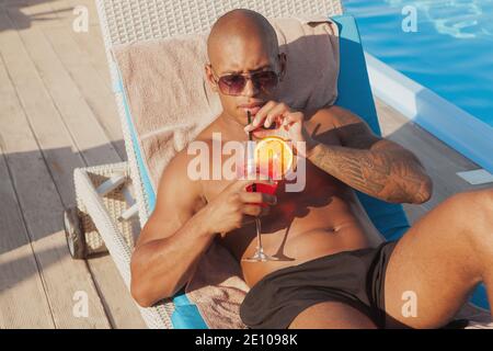 Bell'uomo africano gustando un delizioso cocktail a bordo piscina. Un giovane atletico tatuato uomo che beve un cocktail vicino alla piscina mentre si fa il bagno di sole Foto Stock