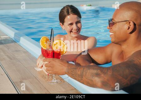 Bella donna felice che ride, parlando con il suo ragazzo mentre beve in piscina. Coppia multirazziale amorevole che celebra la loro estate Foto Stock