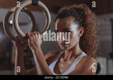 Primo piano di una bella atleta crossfit femminile che guarda alla fotocamera concentrata, tenendo anelli ginnici. Attraente donna atletica che si prepara per h Foto Stock