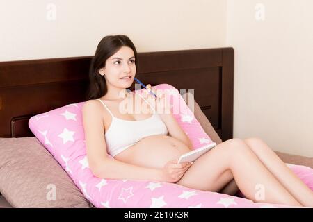 Donna incinta che scrive la lista dei nomi del bambino sul divano, closeup. La mamma incinta scrive in un taccuino e in un elenco di cose da fare shopping. Foto Stock