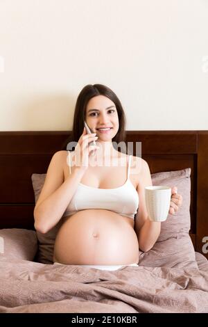 giovane donna incinta seduto a letto e bere caffè e parlare messaggio a casa. concetto di maternità. Foto Stock