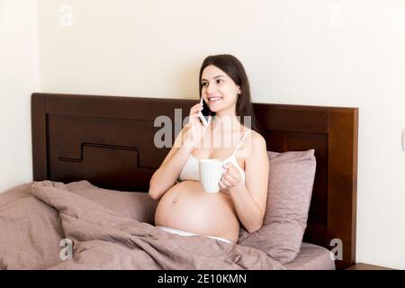 giovane donna incinta seduto a letto e bere caffè e parlare messaggio a casa. concetto di maternità. Foto Stock