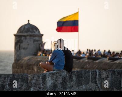 Coppia seduta sulle mura della città con bandiera nazionale colombiana che sventolava In vento tramonto sul Bastione di Santo Domingo fortezza Cartagena De Indias Bolivar Colomb Foto Stock