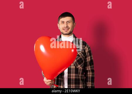 Un ragazzo tiene fuori alla macchina fotografica un palloncino rosso a forma di cuore su uno sfondo rosso. San Valentino. Foto di alta qualità Foto Stock
