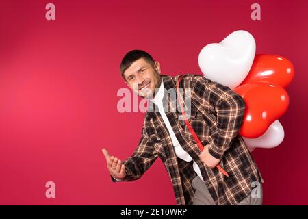Un uomo con un mazzo di palloncini rossi e bianchi che sorridono, che si estende la mano alla telecamera dove sfondo rosso con spazio vuoto. Foto di alta qualità Foto Stock