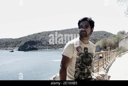 Un uomo etnico indiano del sud asiatico abbronzato con occhiali da sole aviatore, stoppie e T-shirt contro i terreni rocciosi e il mare blu mediterraneo. Ritratto di a. Foto Stock