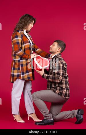San Valentino. Un uomo in ginocchio dà una scatola regalo rossa a forma di cuore. Sfondo rosso. Foto di alta qualità Foto Stock
