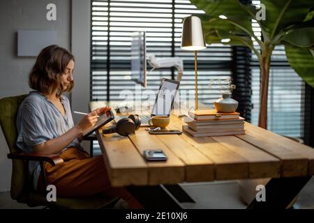 Giovane donna vestita casualmente avendo un po' di lavoro creativo, disegnando su un tablet digitale, seduto nell'accogliente ed elegante ufficio domestico Foto Stock