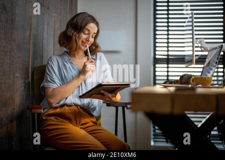 Giovane donna vestita casualmente avendo un po' di lavoro creativo, disegnando su un tablet digitale, seduto nell'accogliente ed elegante ufficio domestico Foto Stock