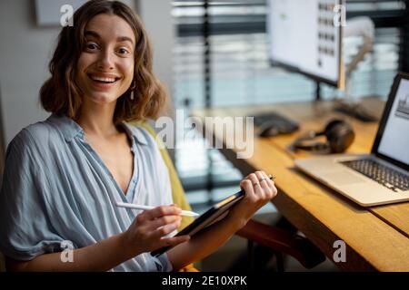 Giovane donna vestita casualmente avendo un po' di lavoro creativo, disegnando su un tablet digitale, seduto nell'accogliente ed elegante ufficio domestico Foto Stock