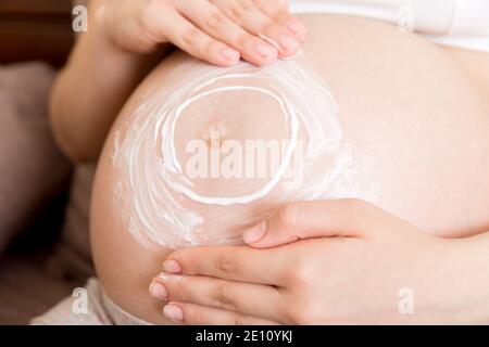 una bambina incinta si siede a casa sul letto e gli spalma una crema anti-stretch sul suo stomaco. Gravidanza, maternità, preparazione e attesa conce Foto Stock