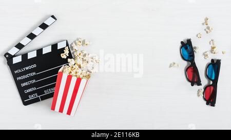 Clapperboard per cinema, occhiali 3d colorati e secchio rosso con popcorn su sfondo bianco in legno. Sfondo cinematografico. Foto Stock