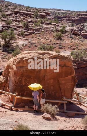 Una coppia sotto un ombrello giallo che guarda l'arte rupestre dei nativi americani sulla Birthing Rock, Kane Creek Road, Moab, Utah, USA. Foto Stock