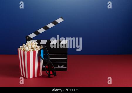 Clapperboard per cinema, occhiali 3d colorati e secchio a righe rosse con popcorn su sfondo rosso e blu. Concetto di cinema. Foto Stock
