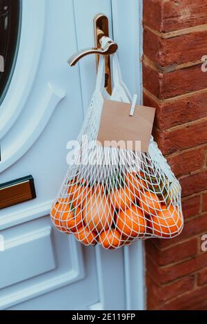 Borsa a corda riutilizzabile in materiali ecologici con agrumi e cartoncino vuoto appeso alla maniglia della porta. Cibo sano contadino locale Foto Stock