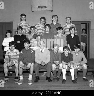 Anni '60, i membri storici, giovani e anziani del Bowill Youth Club, nella loro divisa PE siedono e si levano in piedi insieme, con il leader della squadra del Youth Club nella sala sportiva, per una foto di gruppo, Fife, Scozia, UK. Foto Stock