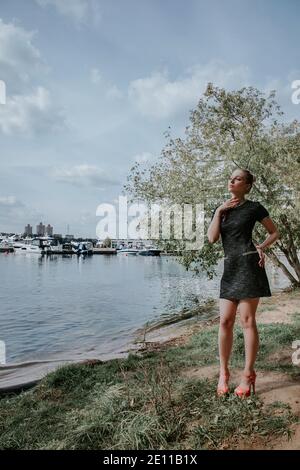 Bella ragazza bionda si trova nel parco vicino all'acqua con le barche sullo sfondo; indossa un abito elegante corto scuro e tacchi alti Foto Stock