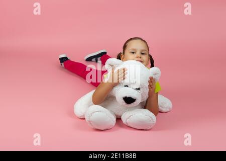 giocosa ragazza contented con i ponytail e pantaloni rossi e luce la t-shirt verde si trova sul pavimento e abbraccia il teddy bianco orso isolato su sfondo rosa Foto Stock