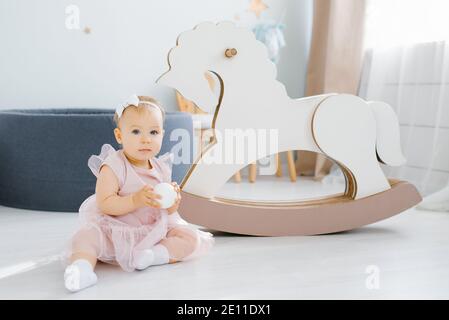 Un bambino di un anno dall'occhio blu carino in un vestito rosa tiene una piccola palla bianca nelle mani e si siede sul pavimento nella stanza dei bambini, con un sw cavallo Foto Stock