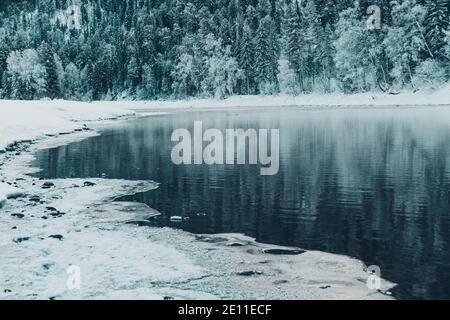 Riva ghiacciata del lago con foresta innevata. Clima freddo, letto del fiume ghiacciato a causa di un brusco scatto freddo Foto Stock