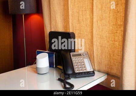 scrivania da ufficio con attivazione vocale, assistente virtuale intelligente, telefono fisso e due router su una scrivania bianca con lampada che si illumina Foto Stock