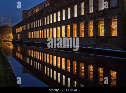 Una sezione di un vecchio mulino, in fase di ristrutturazione, con le finestre illuminate che fanno un disegno lineare nel canale vicino. Foto Stock