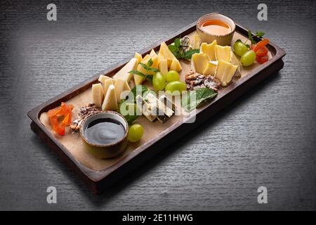 Piatto di formaggi su tavola di legno con uva, menta, miele e marmellata, su sfondo scuro Foto Stock