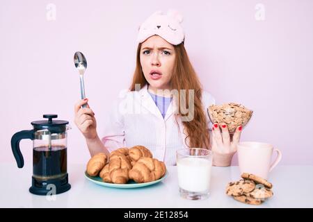Giovane bella donna rossa seduta sul tavolo con la colazione indiziata e confusa espressione. Dubbio concetto. Foto Stock