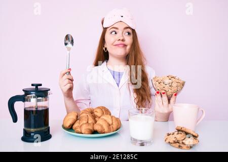 Giovane bella donna rossa seduta sul tavolo con la colazione sorridente guardando da parte e fissando via pensando. Foto Stock