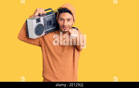 Bell'uomo latino-americano che tiene il boombox, ascoltando la musica che punta a te e alla fotocamera con le dita, sorridendo positivo e allegro Foto Stock