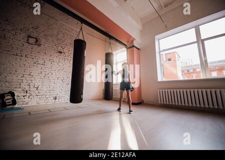 Una bella ragazza kickboxer si sta preparando per le competizioni nella sala di pugilato, praticando la tecnica dei pugni su un sacchetto di punzonatura Foto Stock