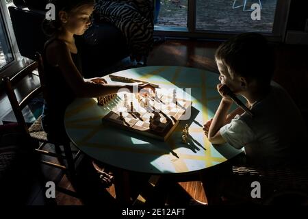 Una giovane ragazza e il suo fratellino giocano a scacchi insieme seduti su un tavolo basso bagnata dalla luce del sole. Foto Stock