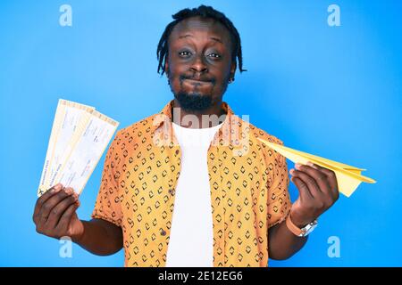 Giovane afroamericano con trecce che tengono l'aereo di carta e carta d'imbarco che soffocano le guance con il viso divertente. Bocca gonfia con aria, che cattura l'aria. Foto Stock