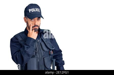 Giovane uomo ispanico che indossa uniforme di polizia che punta all'occhio che ti guarda gesto, espressione sospetta Foto Stock