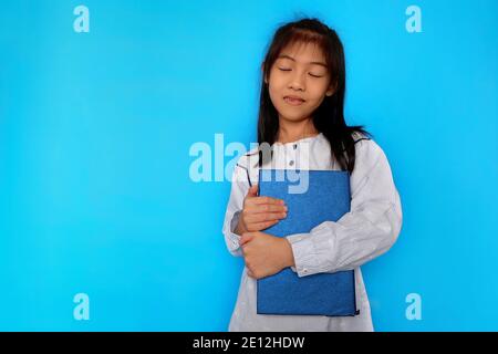 Una giovane ragazza asiatica carina si sente felice dopo aver ricevuto il suo nuovo libro, abbracciandolo stretto e sorridente. Fondo azzurro chiaro. Foto Stock