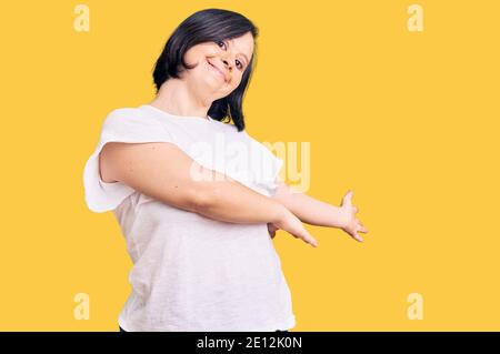 Donna bruna con sindrome di Down indossare casual tshirt bianca invitante per entrare sorridente naturale con la mano aperta Foto Stock