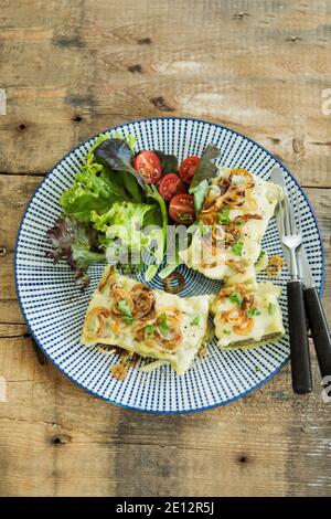 Ravioli svevi con cipolle arrosto e formaggio runny, serviti con insalata su un tavolo di legno giallo Foto Stock