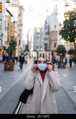 Bella ragazza in abiti alla moda tiene e indossa una maschera usa e getta in strada. Nuovo concetto di stile di vita normale. Foto Stock