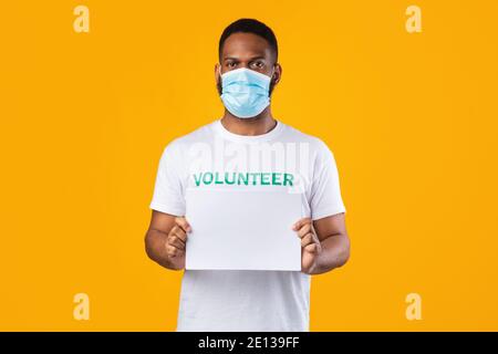 African Volunteer in Mask Holding Blank Poster, Yellow background, Mockup Foto Stock