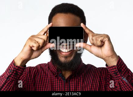 Black Guy copre gli occhi con smartphone che si posa su sfondo bianco Foto Stock