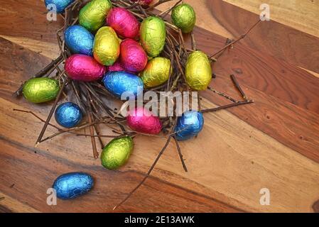 Uova di pasqua colorate avvolte in fogli di alluminio in rosa, verde, blu e giallastro in un nido naturale fatto di bastoni e ramoscelli, contro uno sfondo di legno marrone. Foto Stock