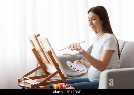 Gravidanza tempo libero. Donna incinta felice pittura sul cavalletto a casa Foto Stock