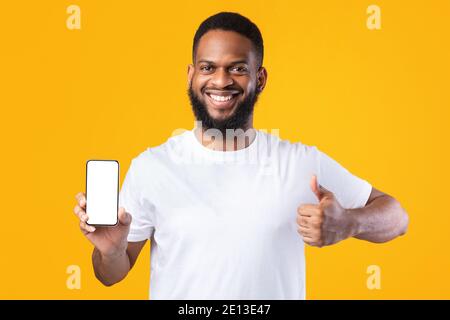 Guy africano che mostra lo smartphone schermo vuoto gesturing Thumbs-Up, sfondo giallo Foto Stock