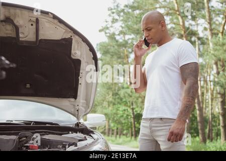 Attraente uomo africano utilizzando smartphone, chiamando assistenza stradale, guardando sotto il cofano della sua auto rotta in autostrada, copia spazio Foto Stock