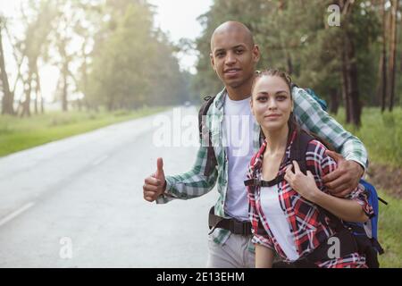 Giovani viaggiatori coppia hitchtrekking sulla strada, spazio copia. Bell'uomo africano e la sua bella ragazza che cattura un'auto sul lato della strada, Foto Stock