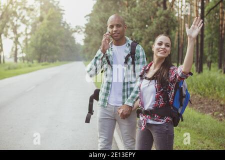 Allegro giovane coppia mista con zaini hitchhiking su strada di campagna, copia spazio. Attraente giovane viaggiatore che agitava per fermare l'auto, indietro Foto Stock