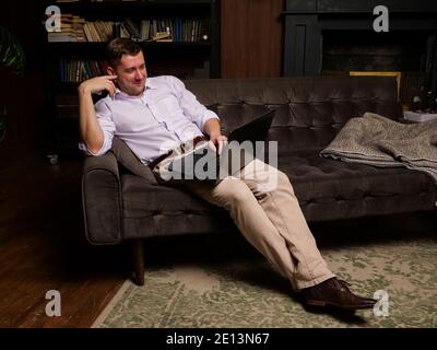 Uomo sorridente che guarda i video sul computer portatile o che parla online mentre si siede sul divano a casa. Foto Stock